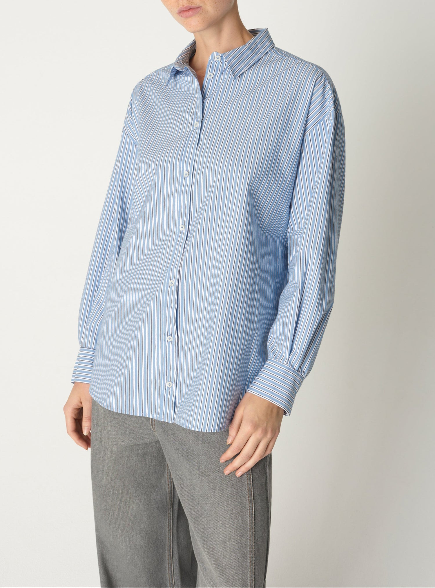 Une personne portant une chemise bleue rayée et un pantalon gris se tient debout sur un fond blanc uni.