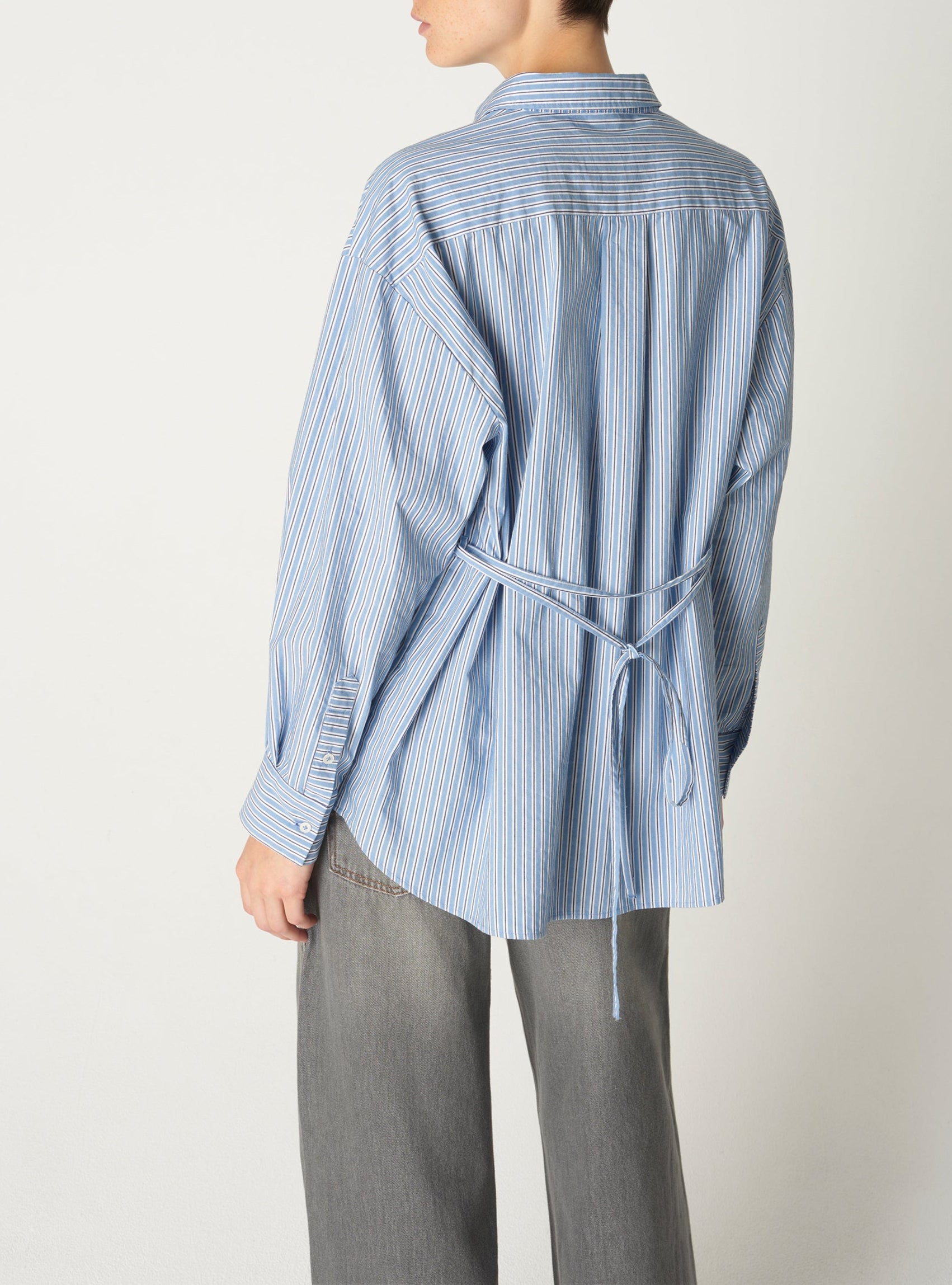 Une personne portant une chemise bleue rayée et un pantalon gris se tient debout sur un fond blanc uni.